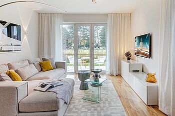 Zu sehen ist ein hell eingerichtetes Wohnzimmer mit bodentiefem Fenster.